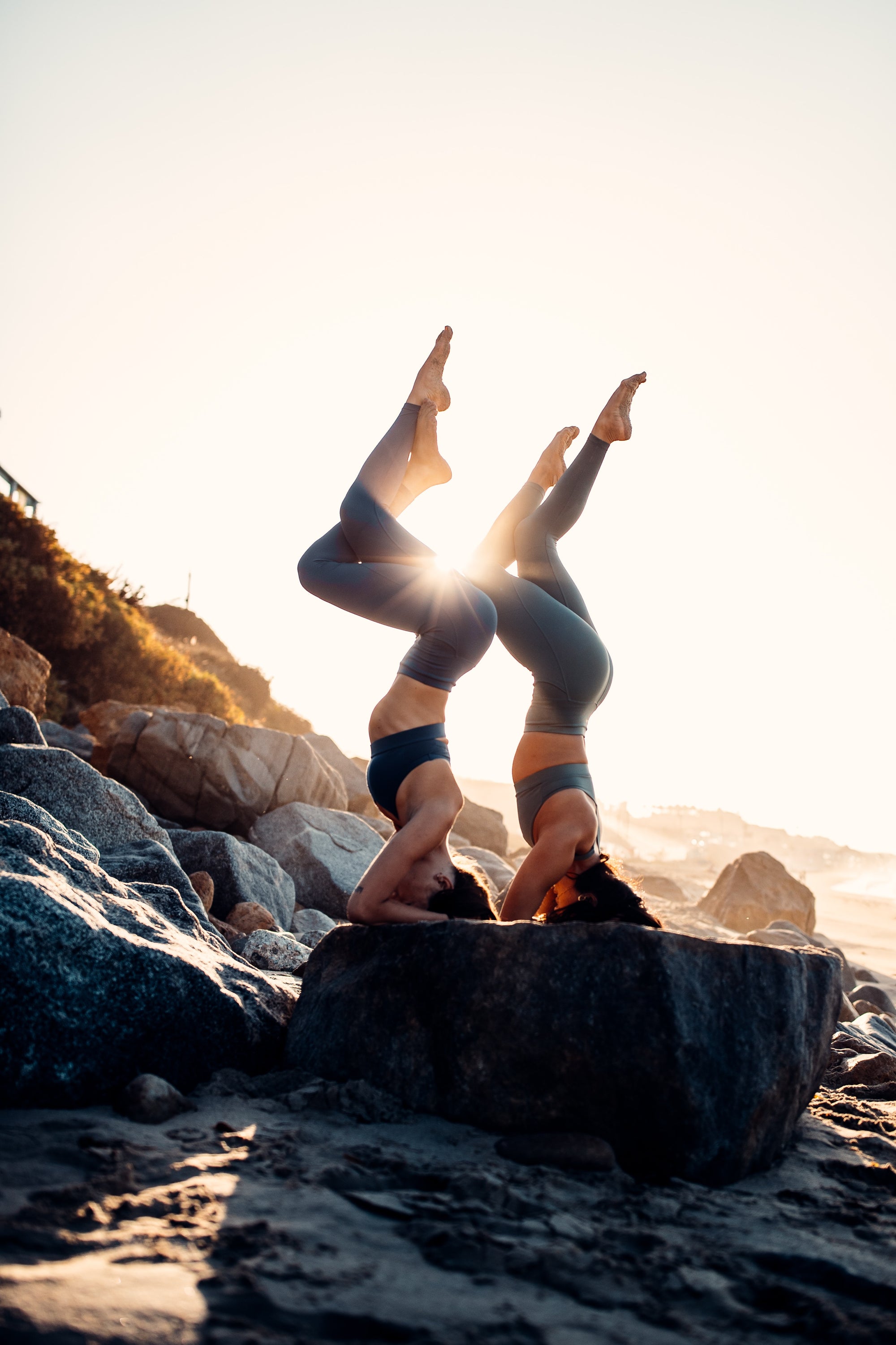 Due donne in posizione di equilibrio yoga con gambe incrociate in verticale, appoggiate su braccia e testa, sole del mattino sullo sfondo